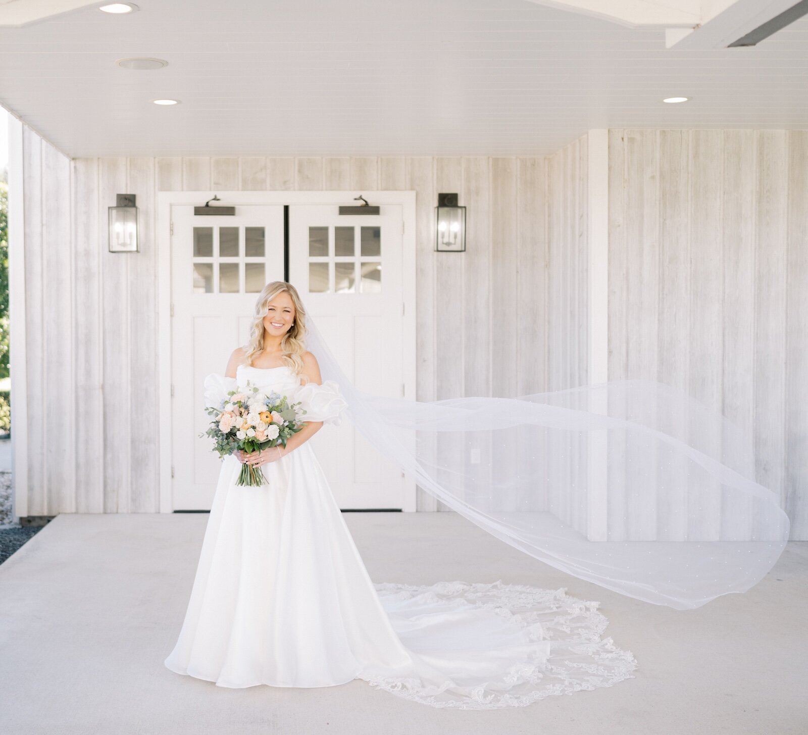 Full-length portrait of a smiling bride in a white gown with puff sleeves and a long, flowing veil standing on a concrete porch in front of a white wood-paneled barn venue entrance.