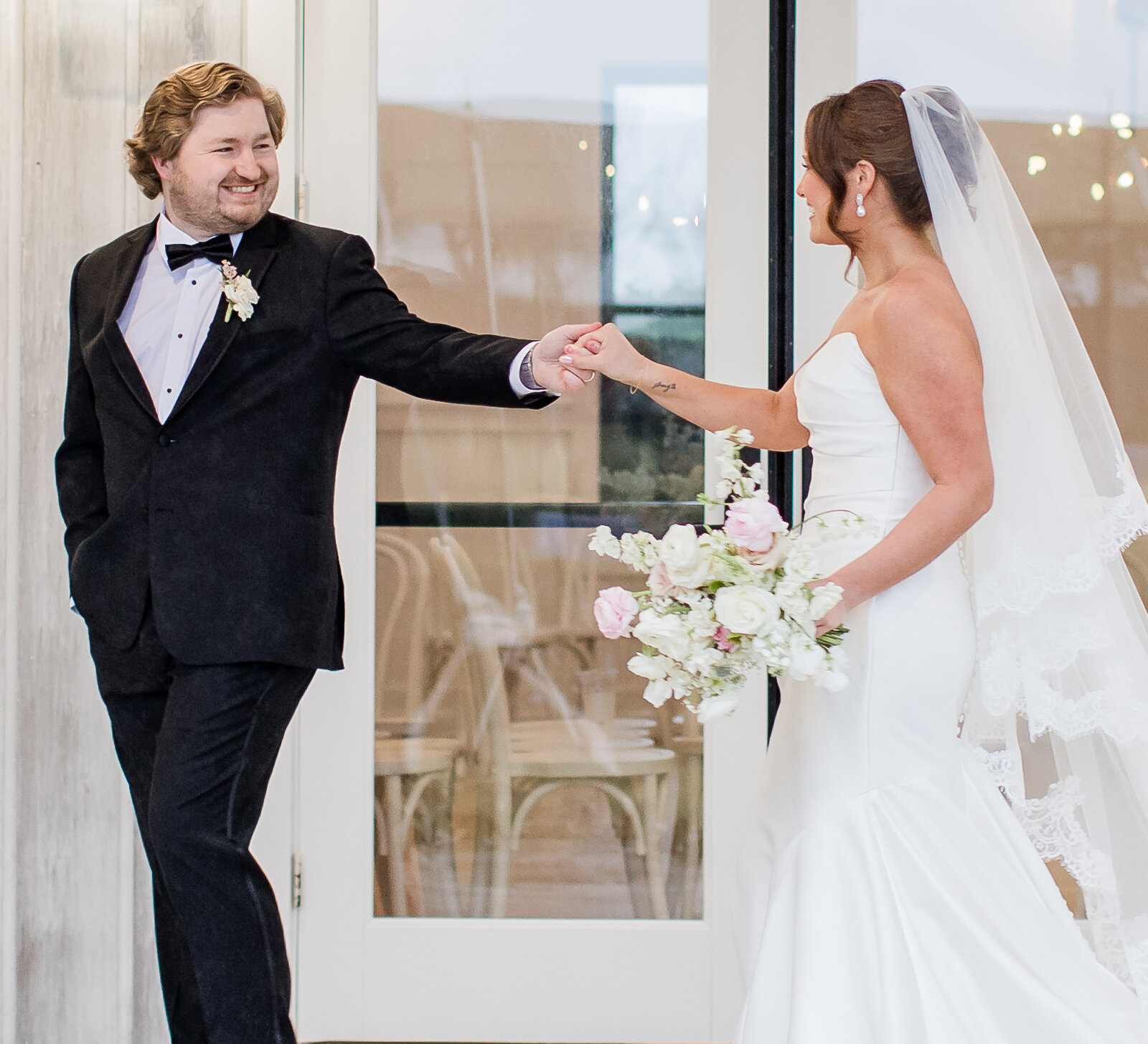 Bride in a strapless white wedding gown and veil holding hands with the smiling groom in a black tuxedo during their first look or portrait session.