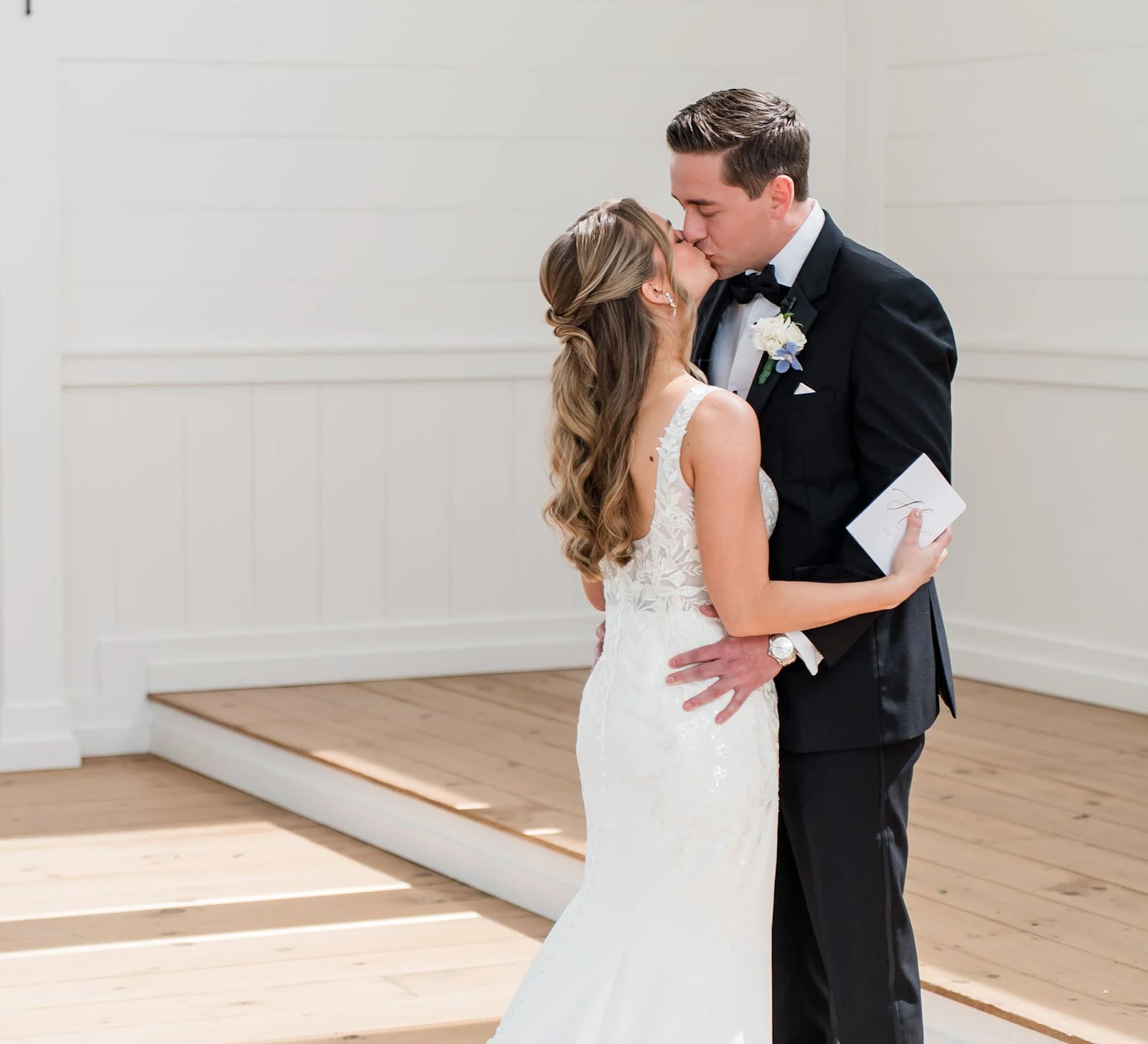 Romantic kiss between the bride in a detailed, white sleeveless gown and the groom in a black tuxedo, holding a small handwritten note on a white wooden porch.