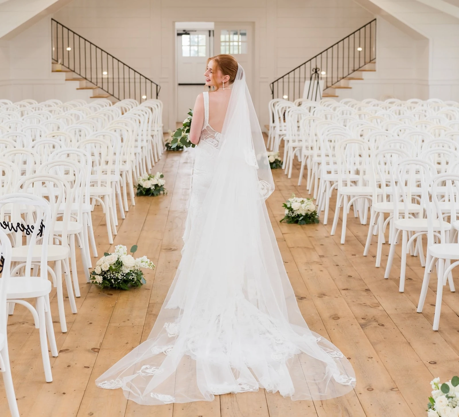 Interior view of a spacious, all-white wedding ceremony hall with vaulted ceilings, natural wood floors, and rows of white chairs. A bride with red hair stands in the center aisle, looking back toward the camera.
