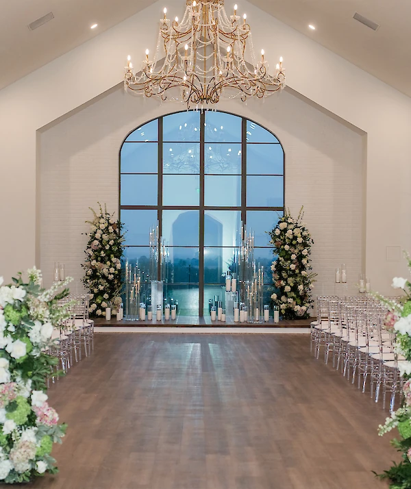 Elegant modern white venue with large arched entryway, tall windows, manicured hedges, and guests gathered near the entrance under a bright blue sky.
