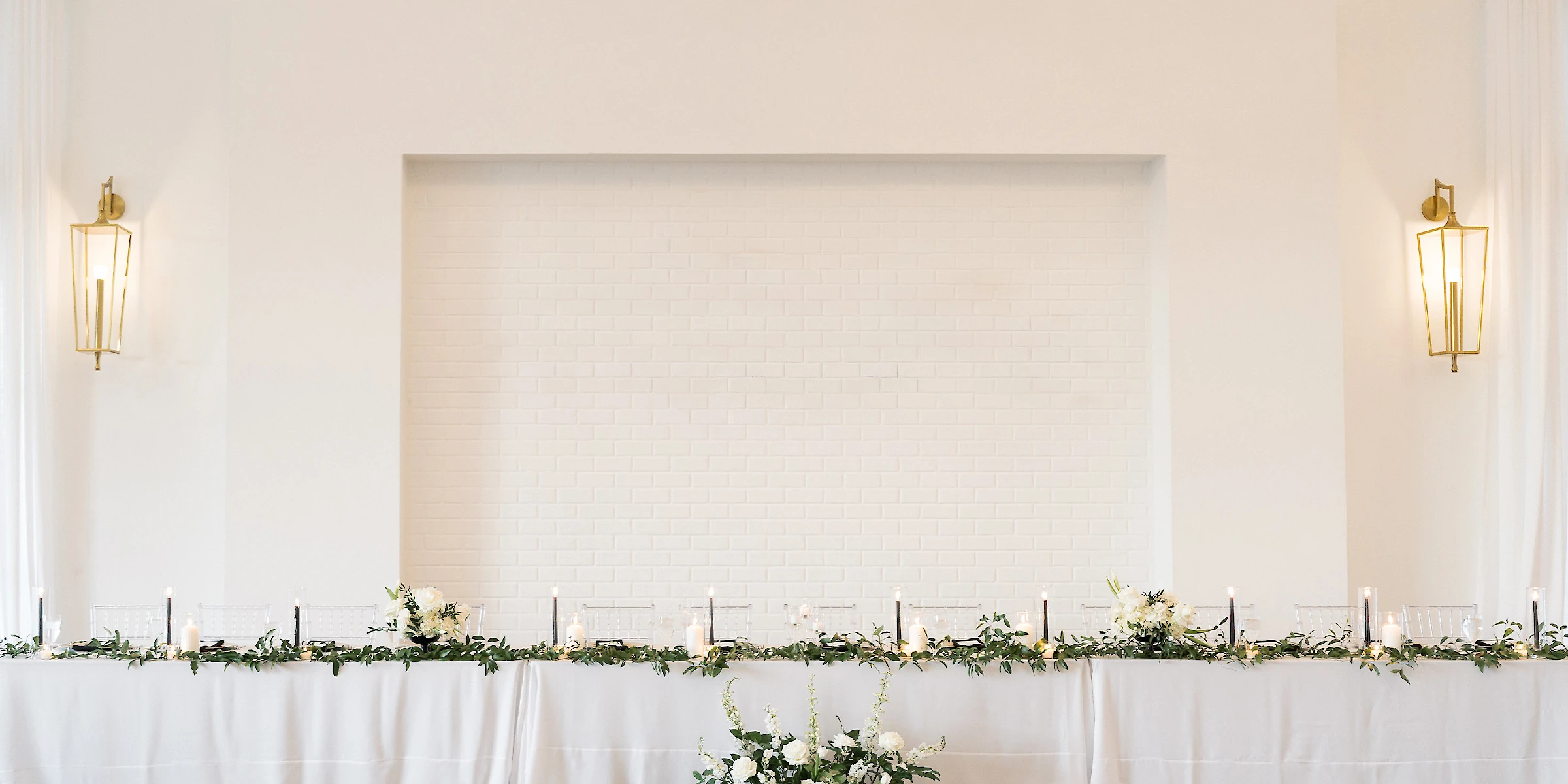 Elegant modern white venue with large arched entryway, tall windows, manicured hedges, and guests gathered near the entrance under a bright blue sky.