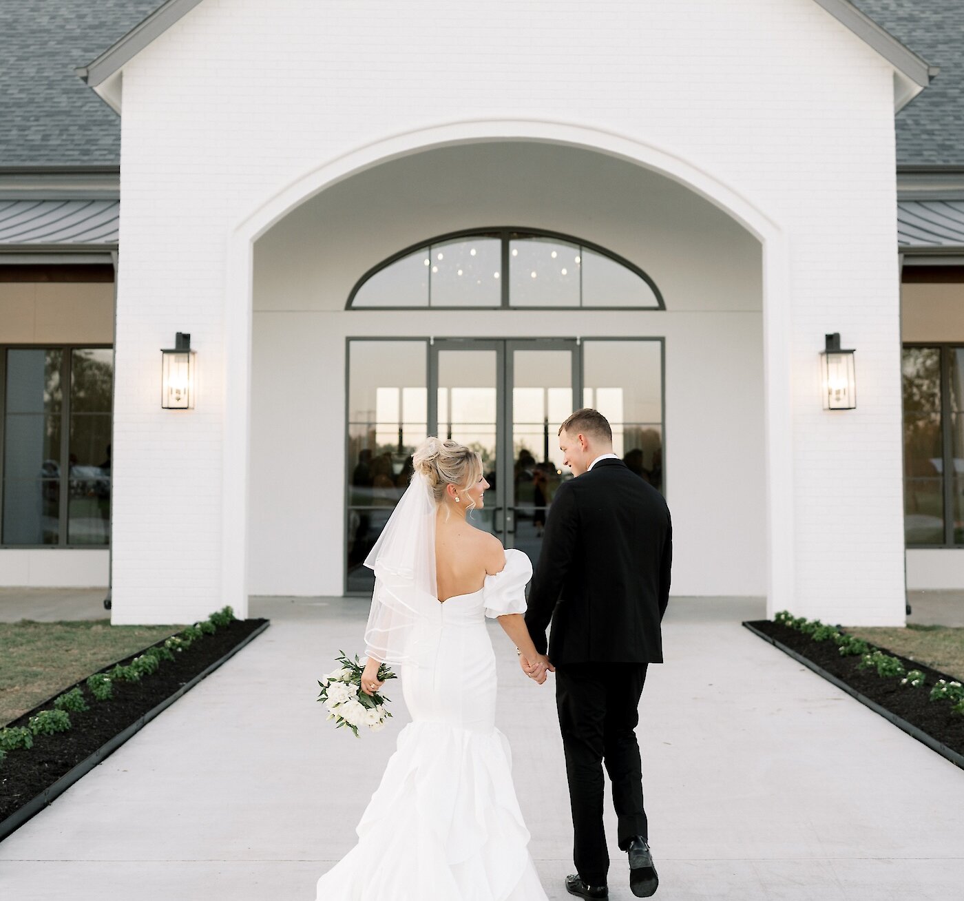 Rear view of a bride and groom holding hands and walking toward the arched entrance of a modern Aylin Acres wedding venue. The bride wears a mermaid-style dress with a dramatic ruffled skirt and an open back.