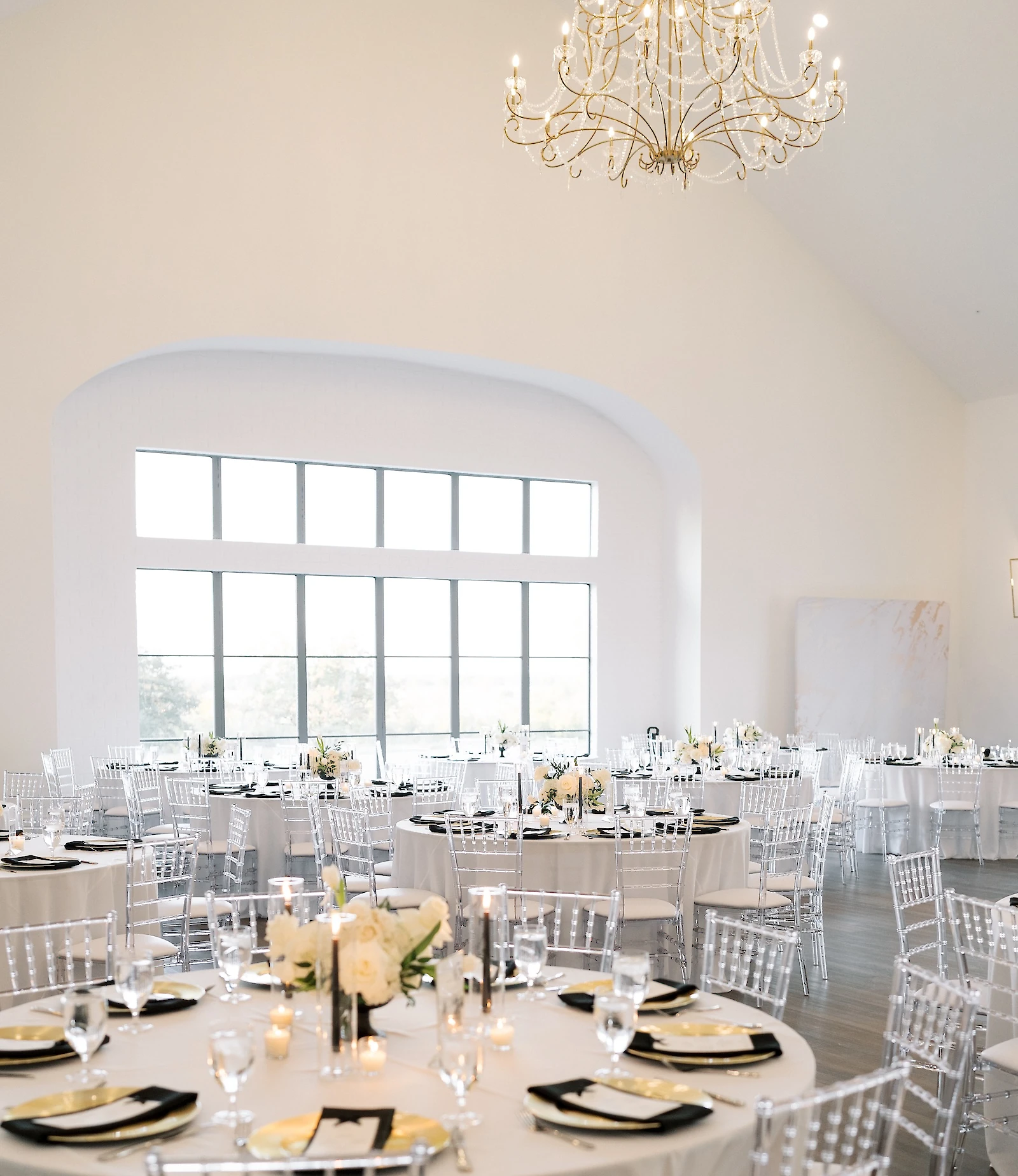 Elegant wedding reception interior with high vaulted ceilings, large arched windows, white round tables set with clear acrylic ghost chairs, and black and gold place settings.