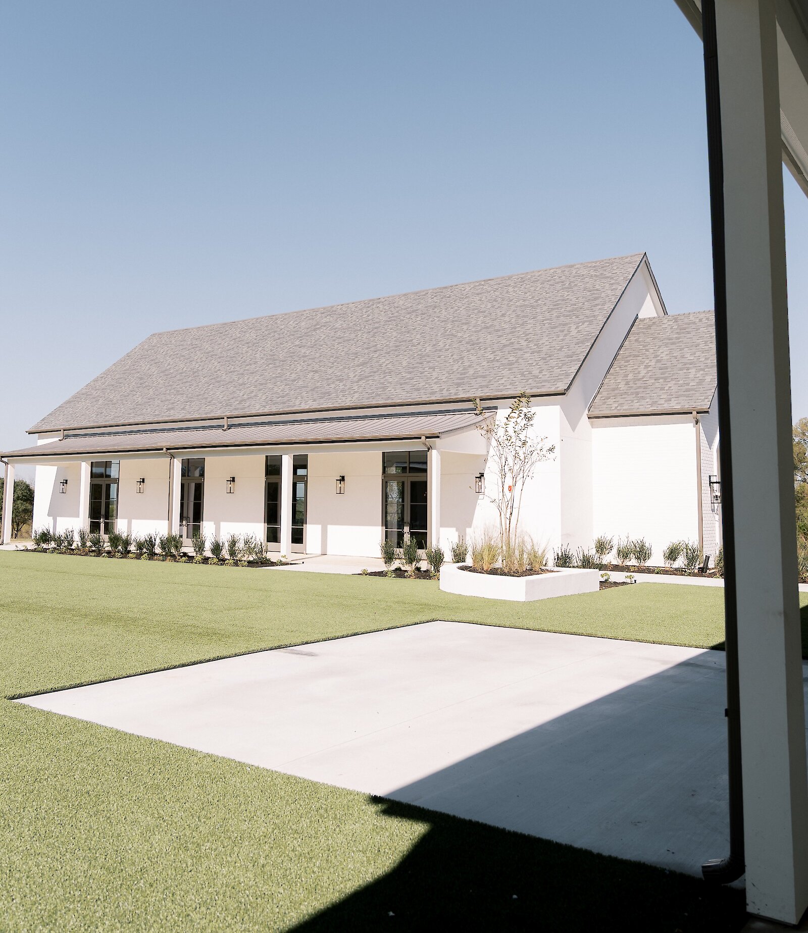 Exterior view of a modern white chapel at Aylin Acres wedding venue with a gray shingled roof, long row of black-framed windows, a covered porch, and a large green lawn with a concrete patio area.