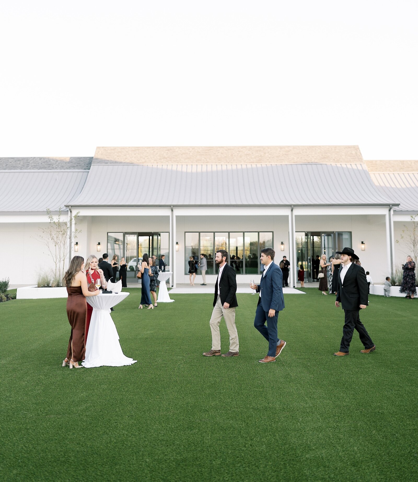 Guests socializing on a manicured green lawn outside a modern white barn venue, set up for a cocktail hour with tall bistro tables and an expansive covered porch in the background.