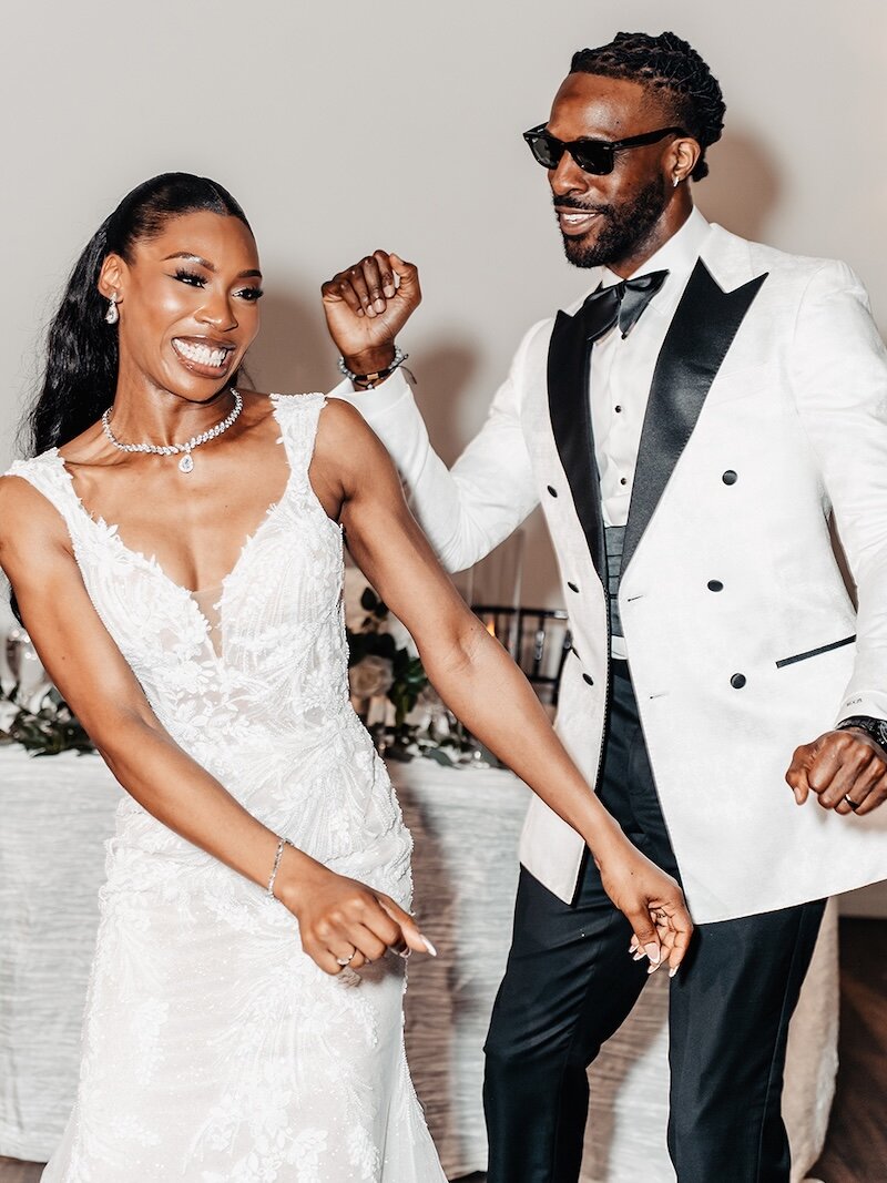Joyful portrait of the bride and groom dancing at their reception. The groom wears a white tuxedo jacket with black lapels and sunglasses, and the bride wears a plunging lace gown with a delicate diamond necklace.