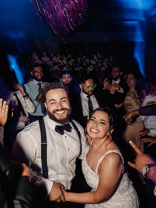 Bride and groom smiling and dancing together on a lively wedding reception dance floor surrounded by guests and colorful lights.