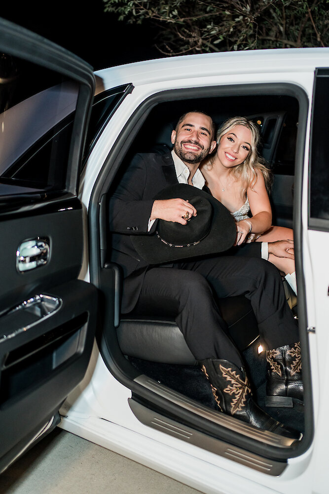 Joyful nighttime photo of the newlyweds seated inside a white luxury getaway car with the door open. The groom smiles and holds a black cowboy hat and wears cowboy boots, while the bride wears a sparkly reception dress.
