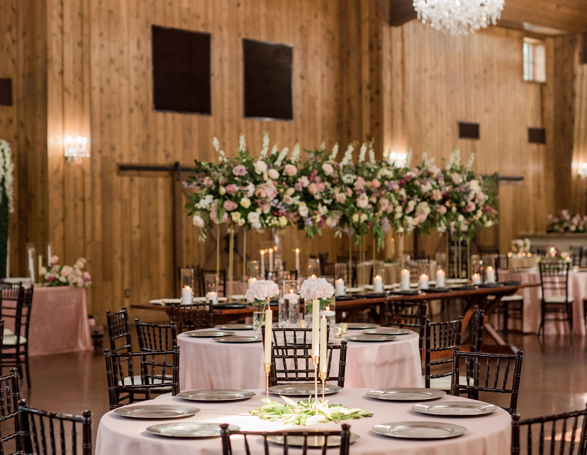 A long wedding reception table featuring bright, colorful floral centerpieces with candles and gold charger plates on a white linen tablecloth.