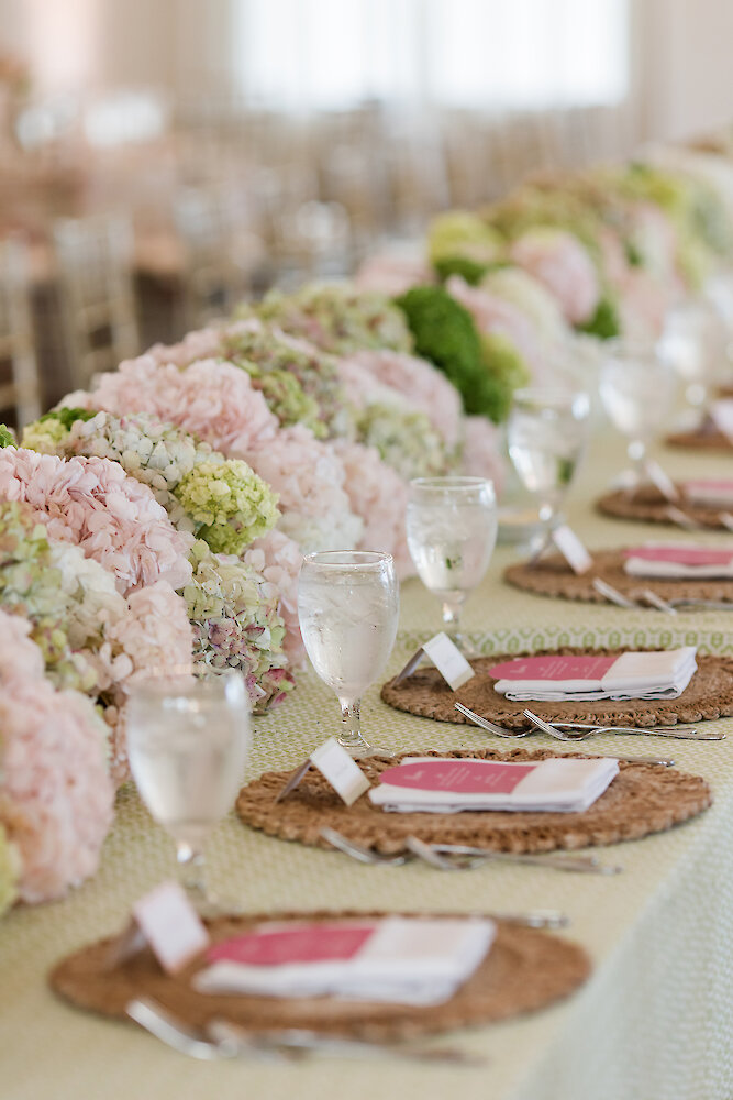 Close-up detail of a wedding reception tablescape featuring a long floral runner composed of blush pink and light green hydrangeas. Place settings include woven placemats, white napkins, and menus with deep pink accents.