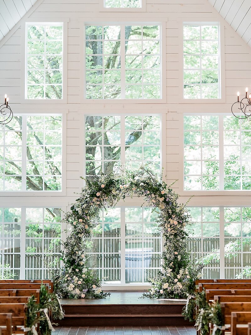 Stunning wedding ceremony altar inside a white wooden chapel with a high vaulted ceiling and a dramatic two-story wall of windows overlooking a lush forest. The altar features a large, asymmetrical floral and greenery arch.