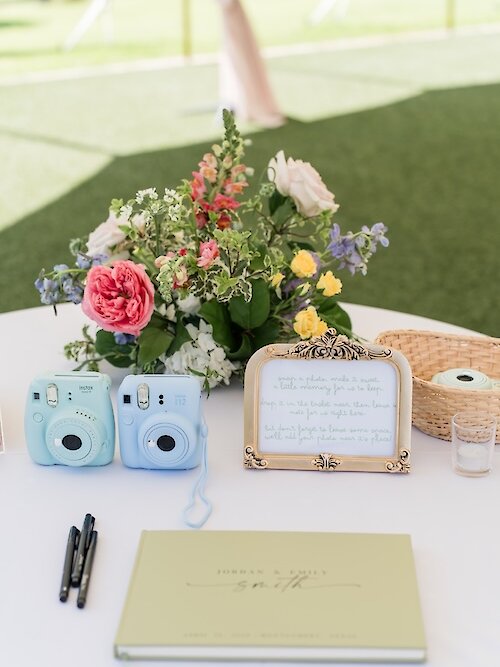 Wedding guest book station detail featuring a light green signature book, two pastel blue instant cameras (Instax), a framed note explaining the photo guest book activity, and a cheerful centerpiece of pink, yellow, and blue flowers.