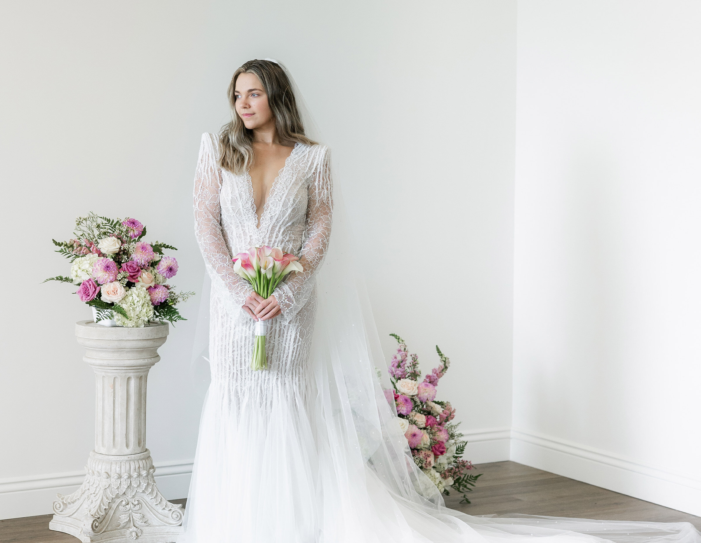 Close-up detail of the bride's shoulder and back showcasing a lace wedding gown with intricate beaded cap sleeves and a sheer veil. The bride holds a delicate bouquet of pink and white calla lilies.