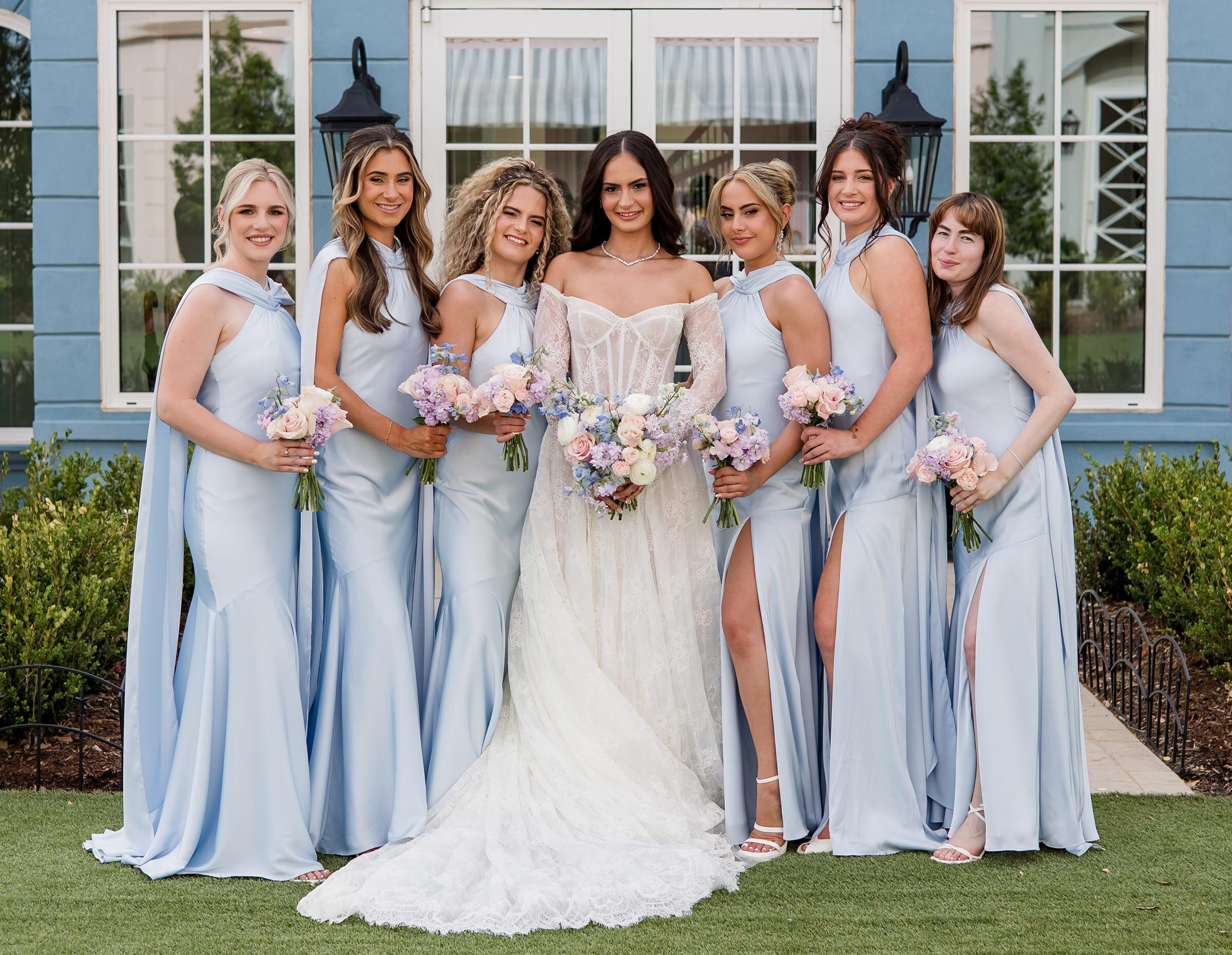 Stylish bridal party portrait of the bride and six bridesmaids standing on a green lawn in front of a light blue building with "THE PLANNING STUDIO" signage. The bridesmaids wear matching light blue satin gowns with high slits, and the bride holds a massive pastel bouquet.