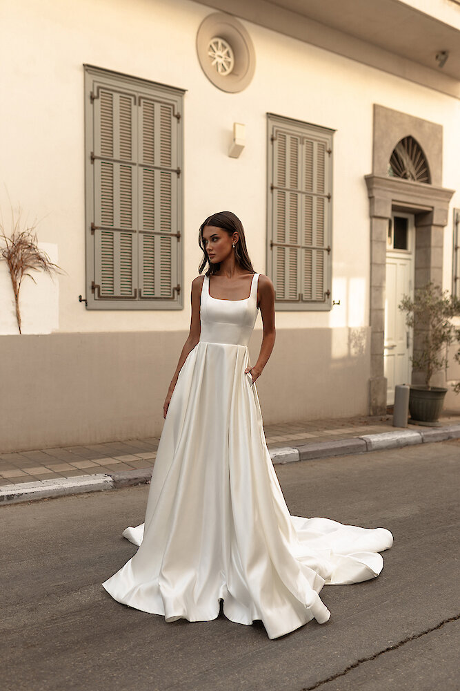 Classic bridal portrait of a model wearing a simple, elegant square-neck satin ballgown with a long train and pockets. She is posing on a European street with a light-colored building facade and shuttered windows in the background.