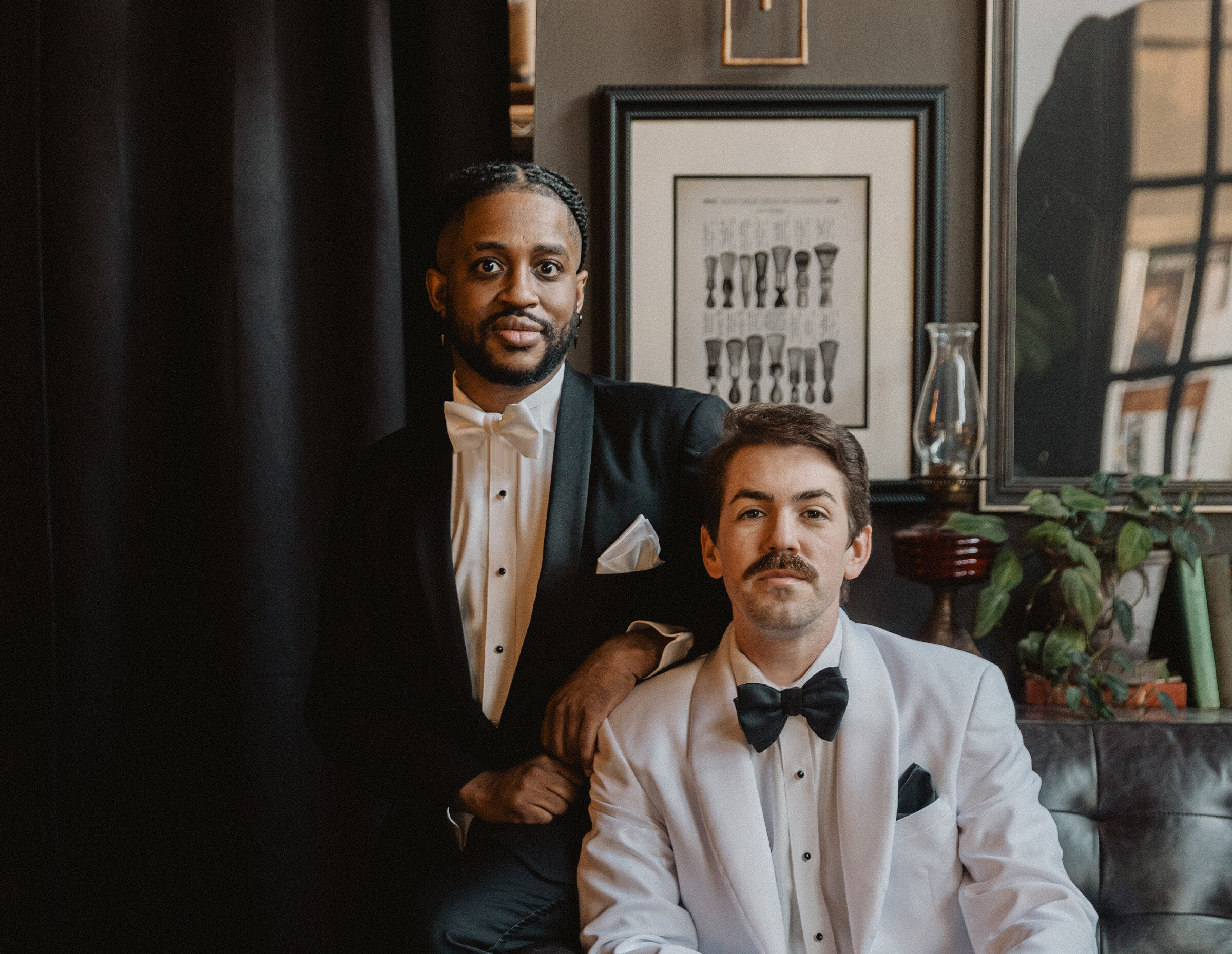 Sophisticated portrait of two men in black tie attire in a dark, moody lounge setting. One man is seated wearing a white tuxedo jacket and the other stands behind him in a black tuxedo jacket, with a black curtain backdrop.