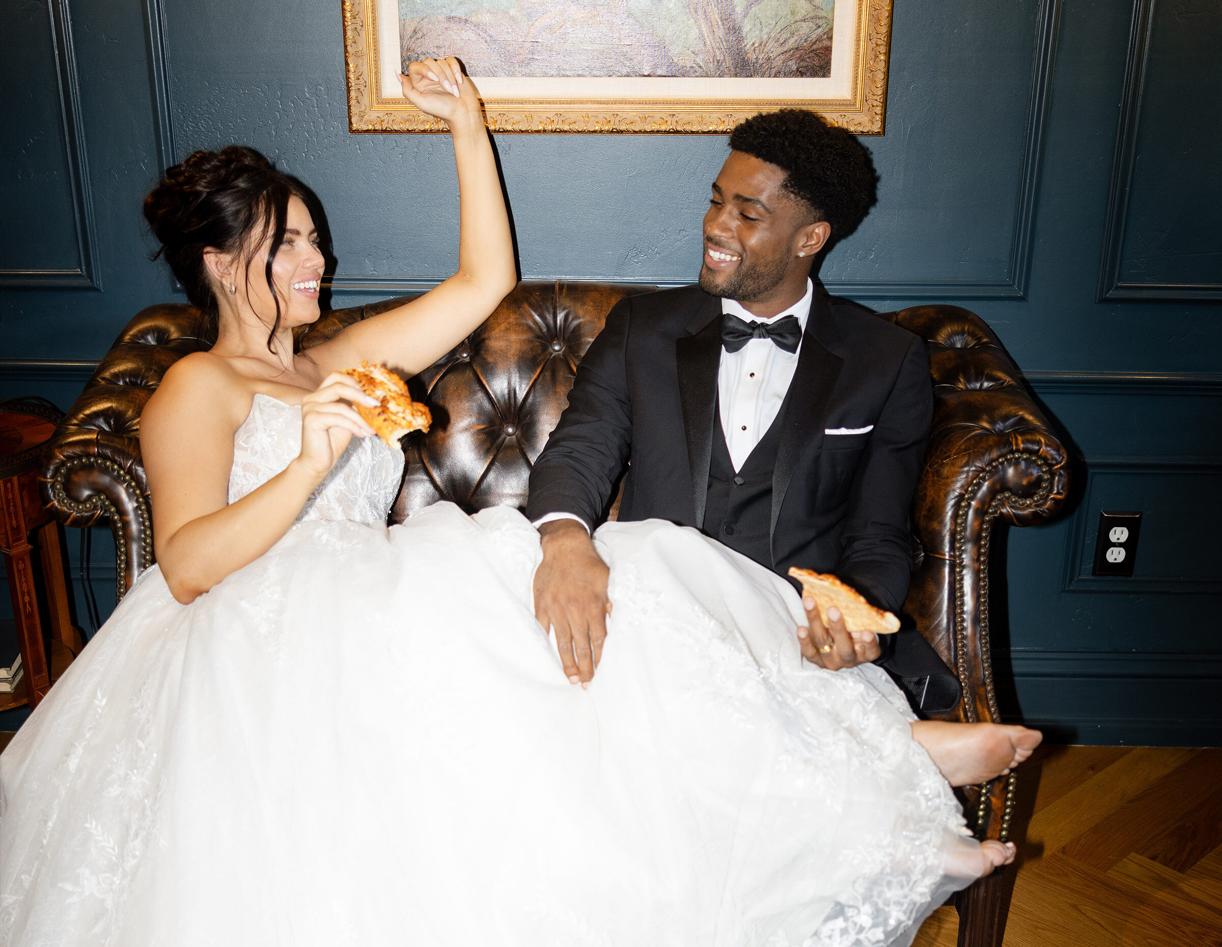 Classic wedding portrait of the couple embracing in front of a dense green ivy wall. The groom wears a blue three-piece suit with a beige tie, and the bride wears a sleek strapless satin gown and holds a pastel mixed floral bouquet.