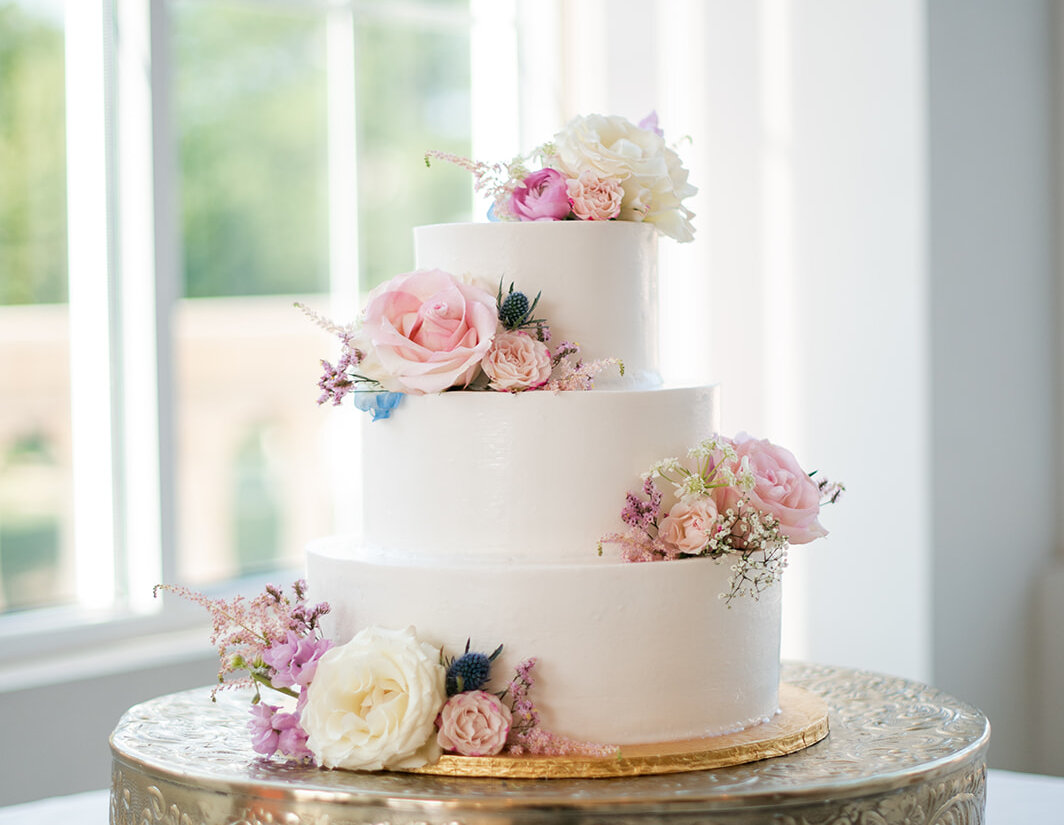 Three-tier white wedding cake decorated with pink and white fresh flowers on an ornate gold cake stand near a bright window.