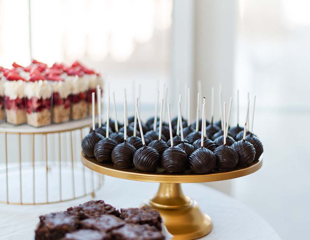 Assorted wedding desserts including chocolate-covered cake pops, layered strawberry parfaits, and stacked brownies displayed on elegant gold and white stands