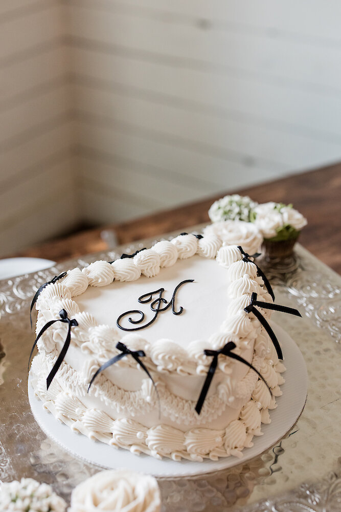 Heart-shaped white wedding cake decorated with black ribbon bows and monogram detail on a clear glass cake stand.