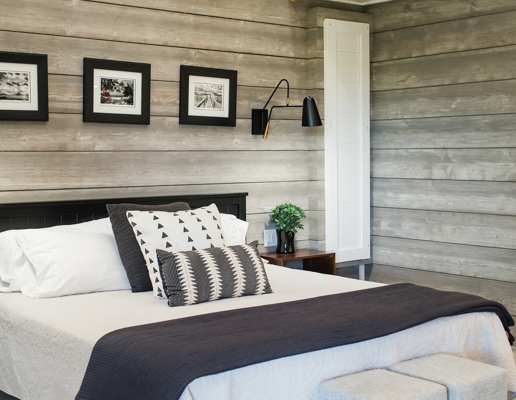 A modern farmhouse bedroom featuring a bed with neutral bedding, a shiplap wood-paneled wall, black framed art, and a small loft balcony.