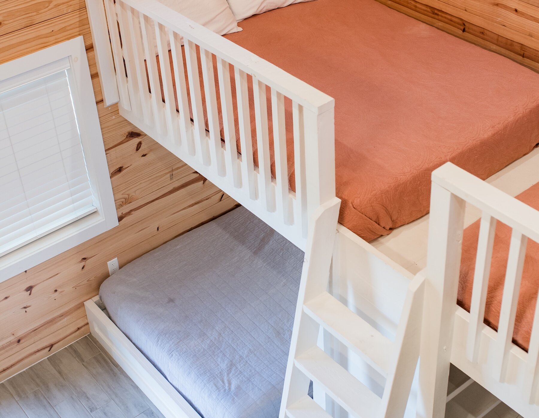 An overhead view of built-in wooden bunk beds with white railings and ladder, featuring a rust-colored top mattress and a light grey bottom mattress.