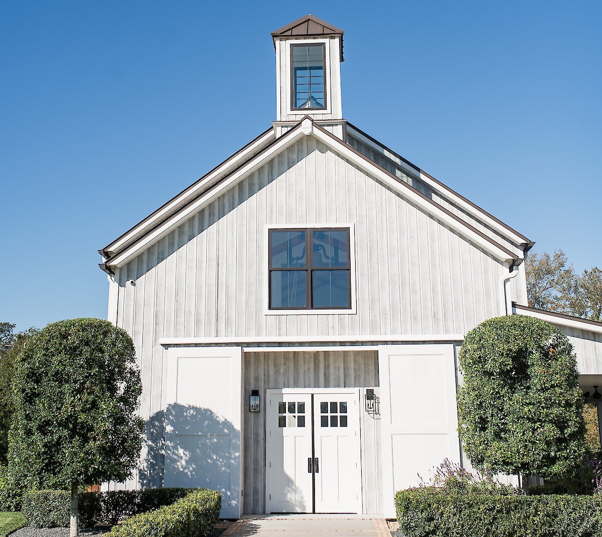 The front view of the weathered white barn at Arrowhead Hill, featuring a center cupola and double white entry doors framed by landscaping.