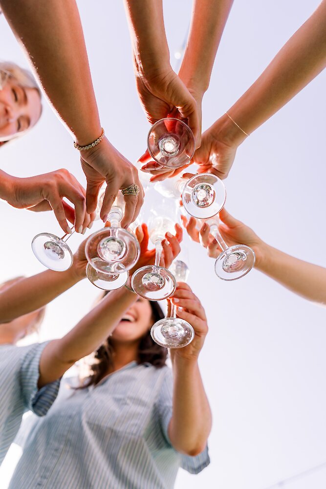A low-angle shot of several women cheering and toasting with empty wine glasses held high against a bright sky.