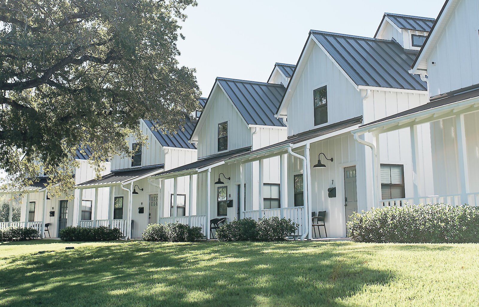 A sunlit row of white modern farmhouse cottages with dark metal roofs, covered porches, and a green lawn in the foreground.