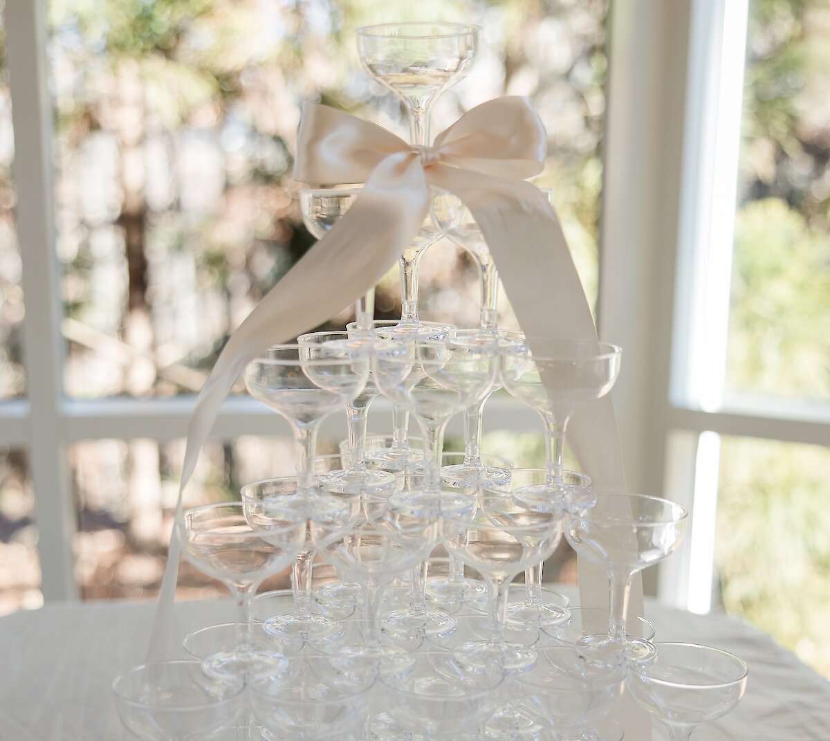 A tiered champagne tower made of clear coupe glasses, decorated with a large, creamy white satin ribbon on top.