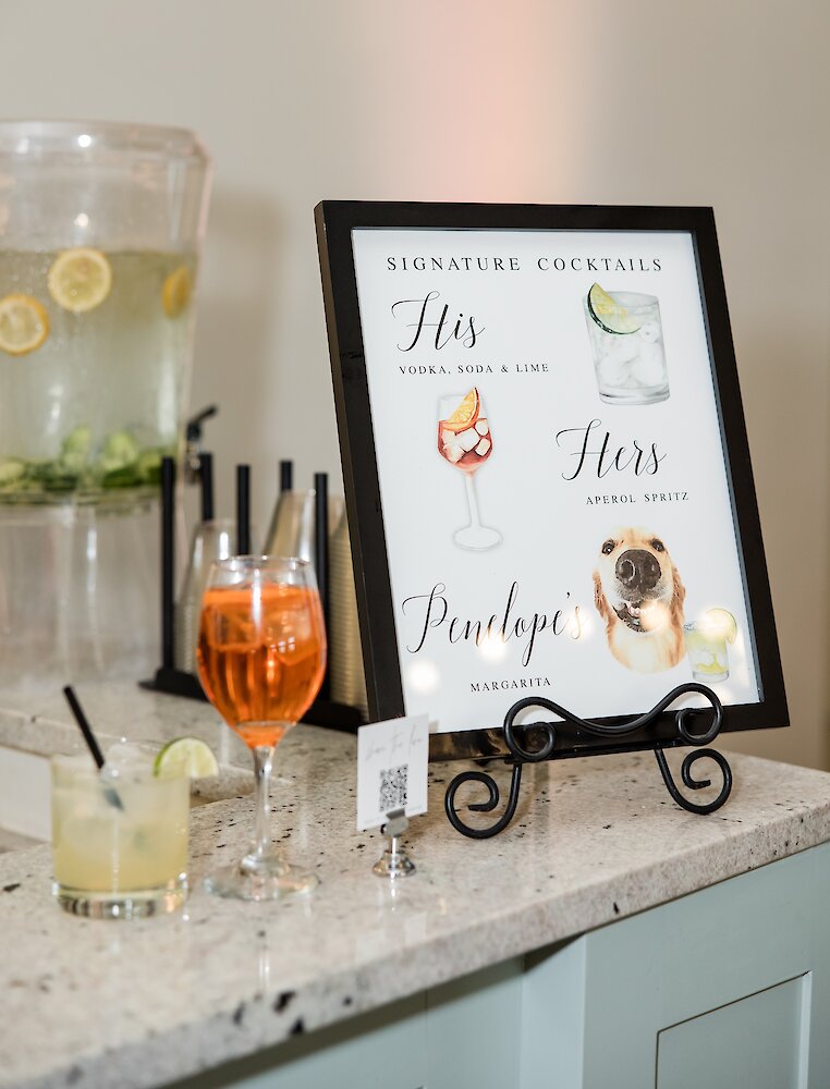 A wedding bar features a sign detailing "His," "Hers," and "Penelope's" signature cocktails with watercolor illustrations, including a dog.