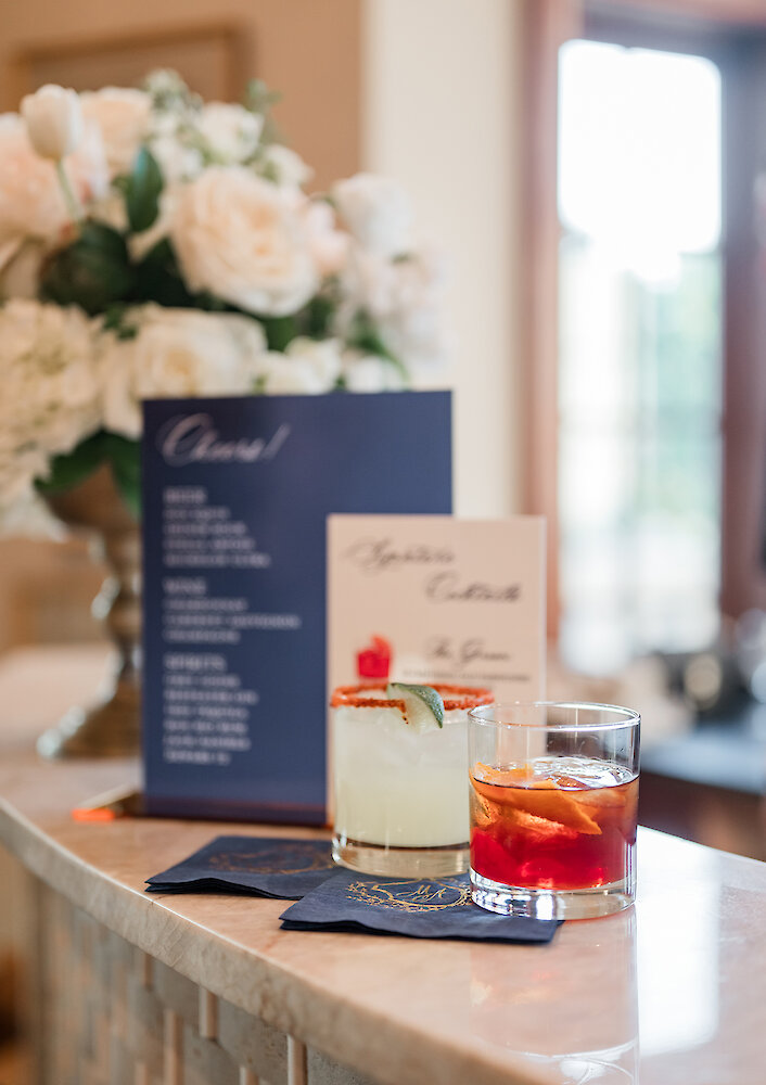 Two colorful cocktails served on a woven placemat, with a pink arched menu and a blurred floral background.