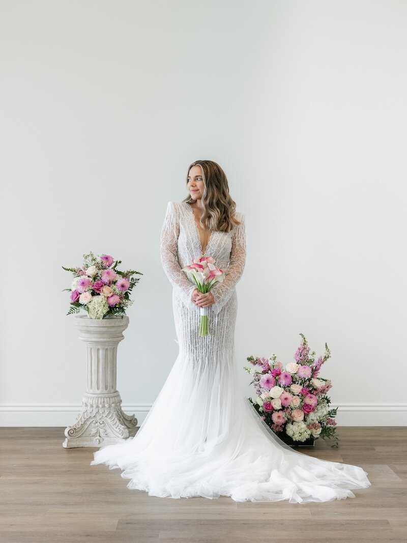 A bride wearing a shimmering, beaded long-sleeve gown stands between two large pink and white floral arrangements.