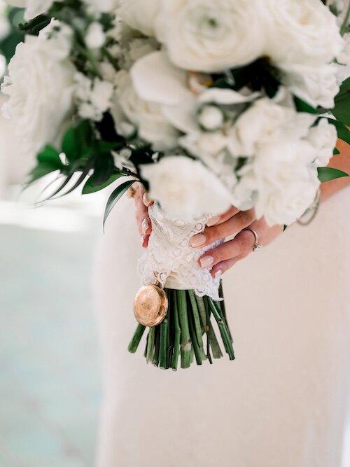 The bride holds her white floral bouquet wrapped in lace, with a sentimental gold pocket watch locket attached to the stems.