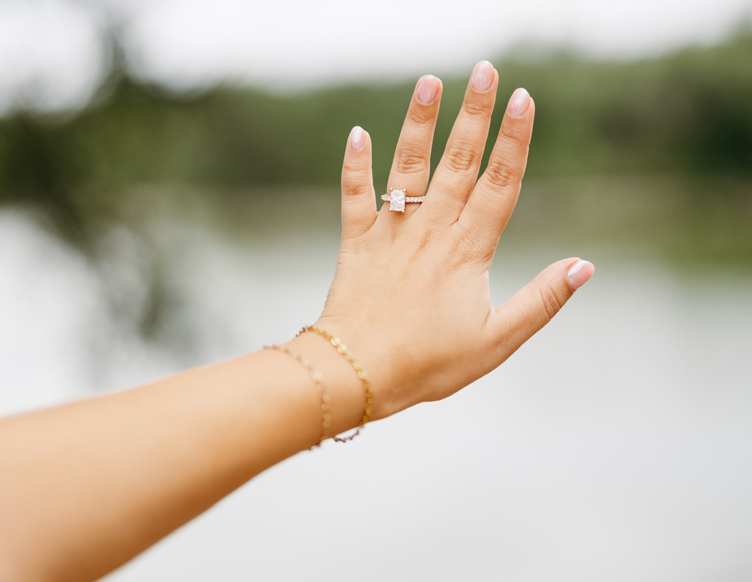 Close-up photo of an engagement ring featuring a large, horizontal emerald-cut diamond with side stones, stacked with a diamond wedding band, worn on the hand of a blonde woman wearing a white knit sweater.