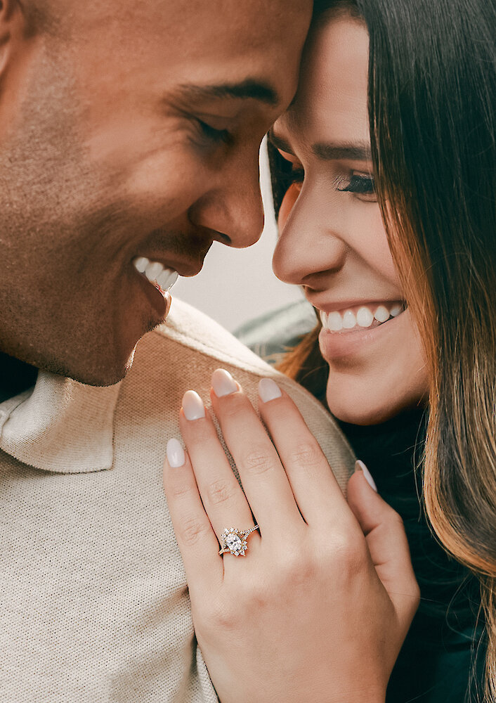 Romantic proposal photo showing a man kneeling on one knee, holding an open ring box to a surprised woman, inside a white wooden gazebo overlooking a misty lake. The woman is wearing a flowing emerald green dress.