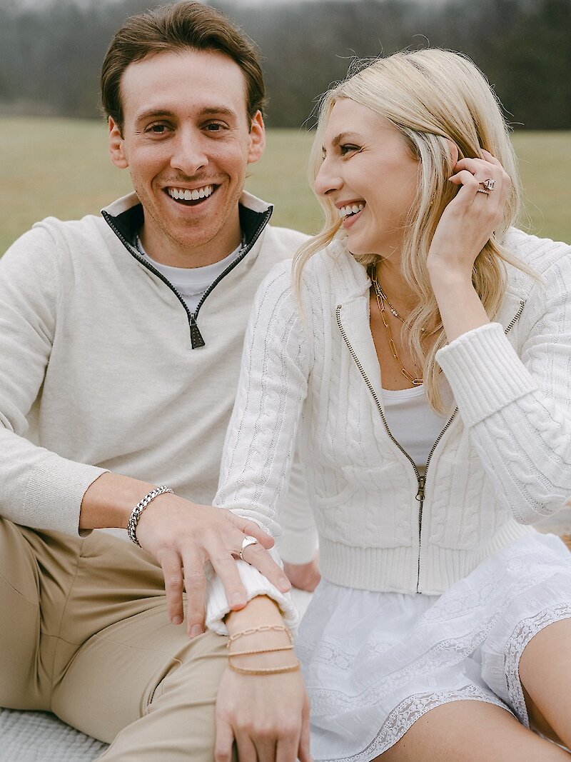 Cheerful engagement portrait of a couple sitting close together outdoors, laughing and smiling. The woman wears a white cable-knit jacket and skirt, and the man wears an off-white half-zip sweater, both showing off their hand jewelry.