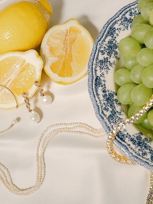 Luxurious still life photo featuring pearl jewelry (necklace, bracelet, and drop earrings) arranged on a white satin background next to bright yellow lemons and green grapes served on a blue and white patterned plate.