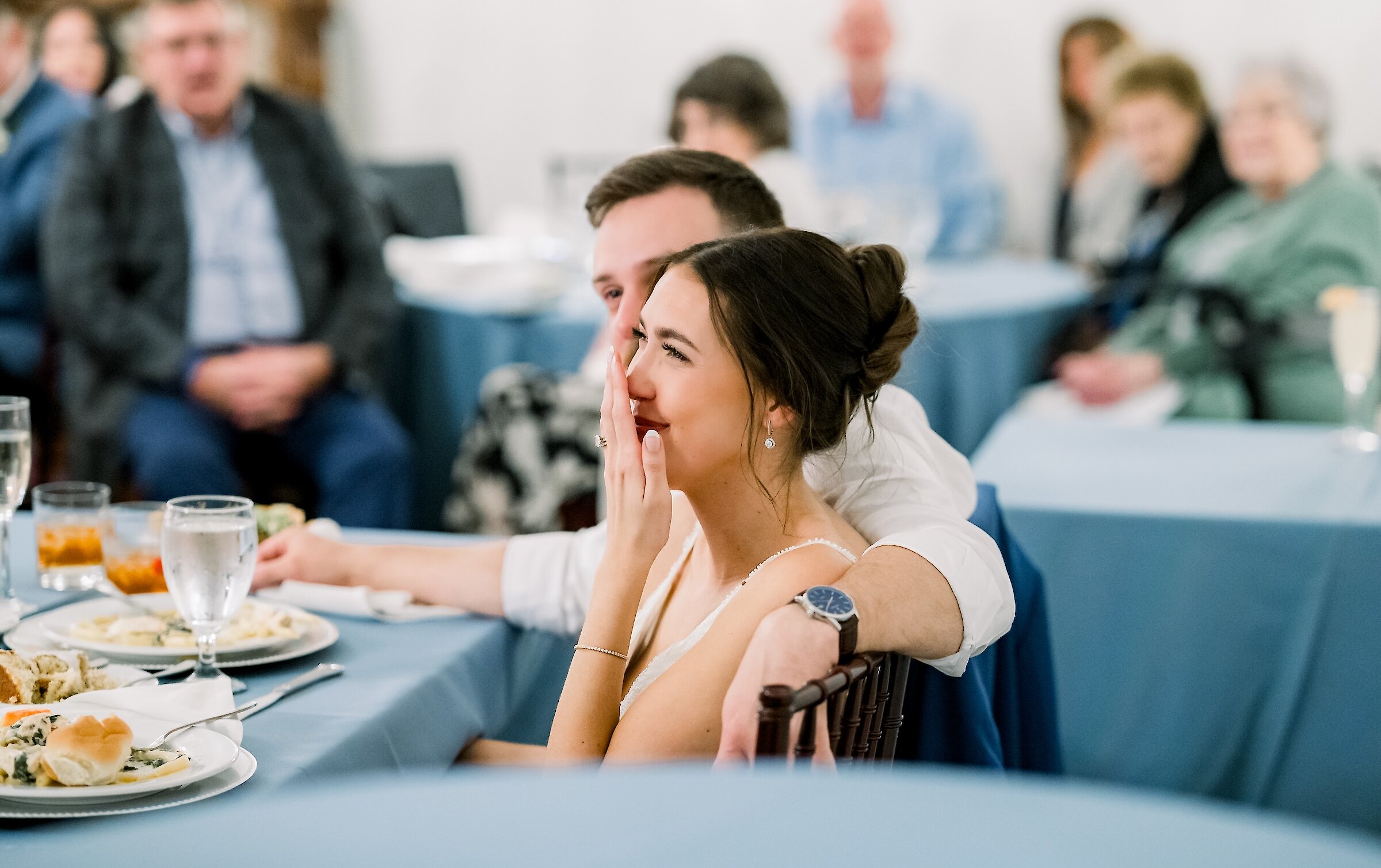 The bride smiles with her hand to her mouth, sitting closely with the groom at a blue-linen covered reception table.