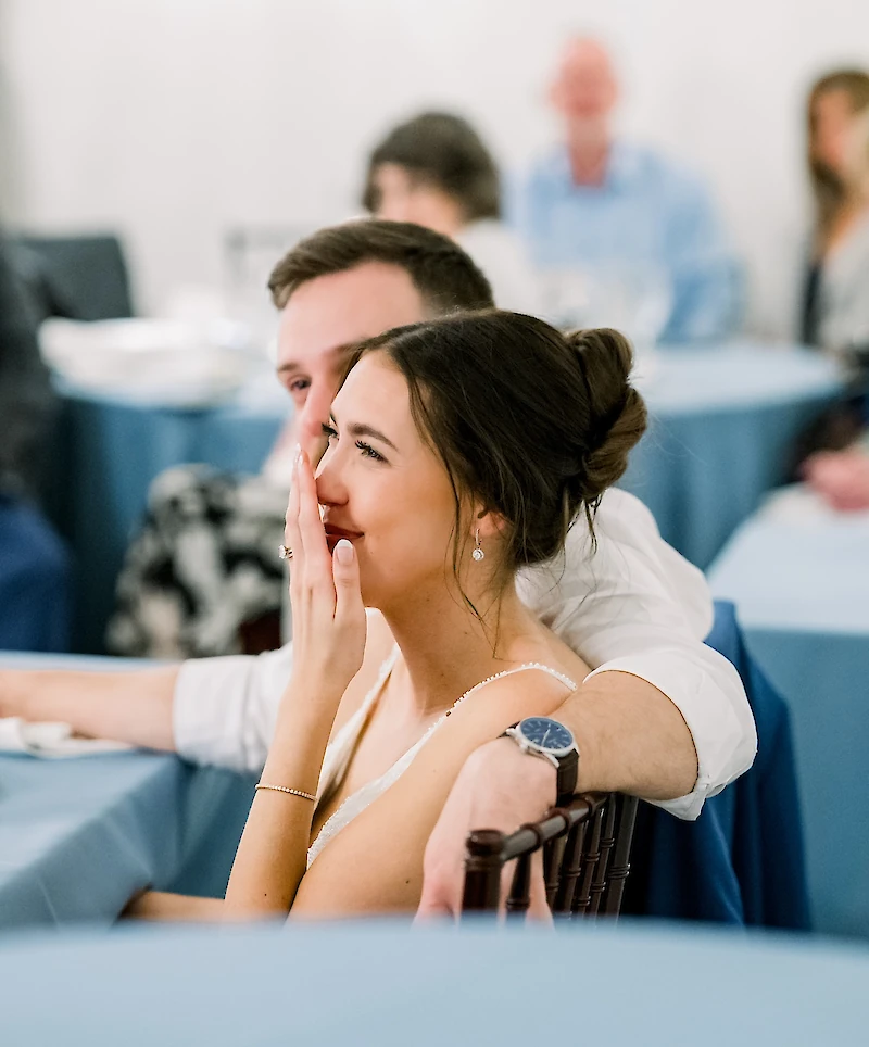The bride smiles with her hand to her mouth, sitting closely with the groom at a blue-linen covered reception table.