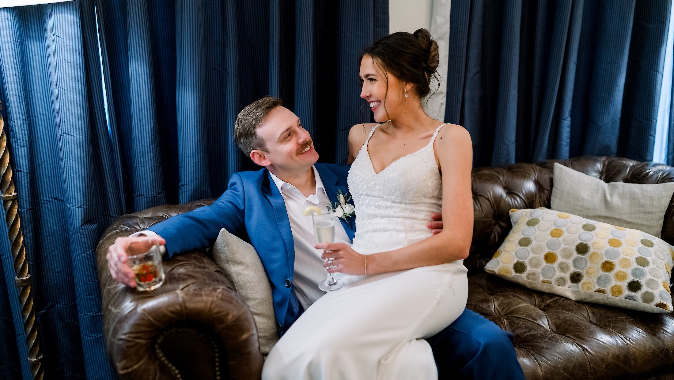 The bride and groom are sitting together on a leather sofa with blue curtains in the background, smiling and holding celebratory drinks.
