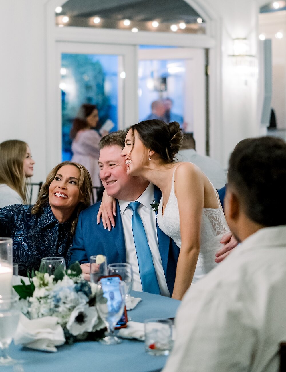 The smiling bride embraces a man, likely her father, at a reception table decorated with blue and white flowers and a blue tablecloth.