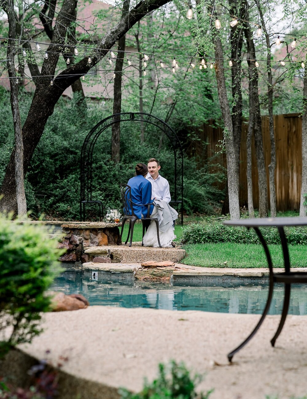 The bride and groom sit on chairs next to a pool, surrounded by a lush forest and string lights hanging from the trees.
