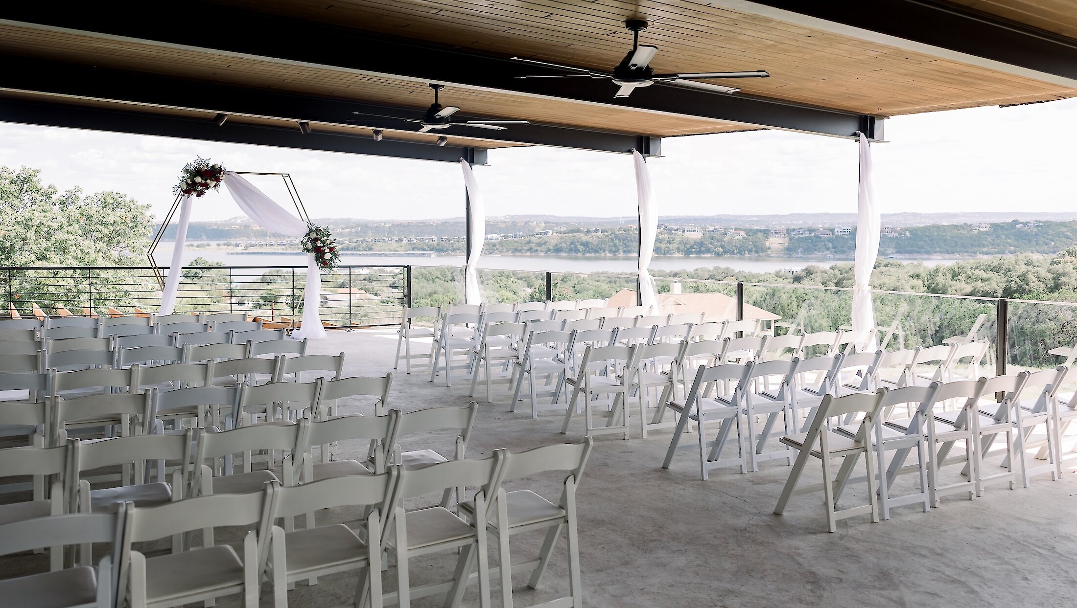 An outdoor covered ceremony space with rows of white folding chairs, a geometric arch, and a panoramic view of the lake and hills.
