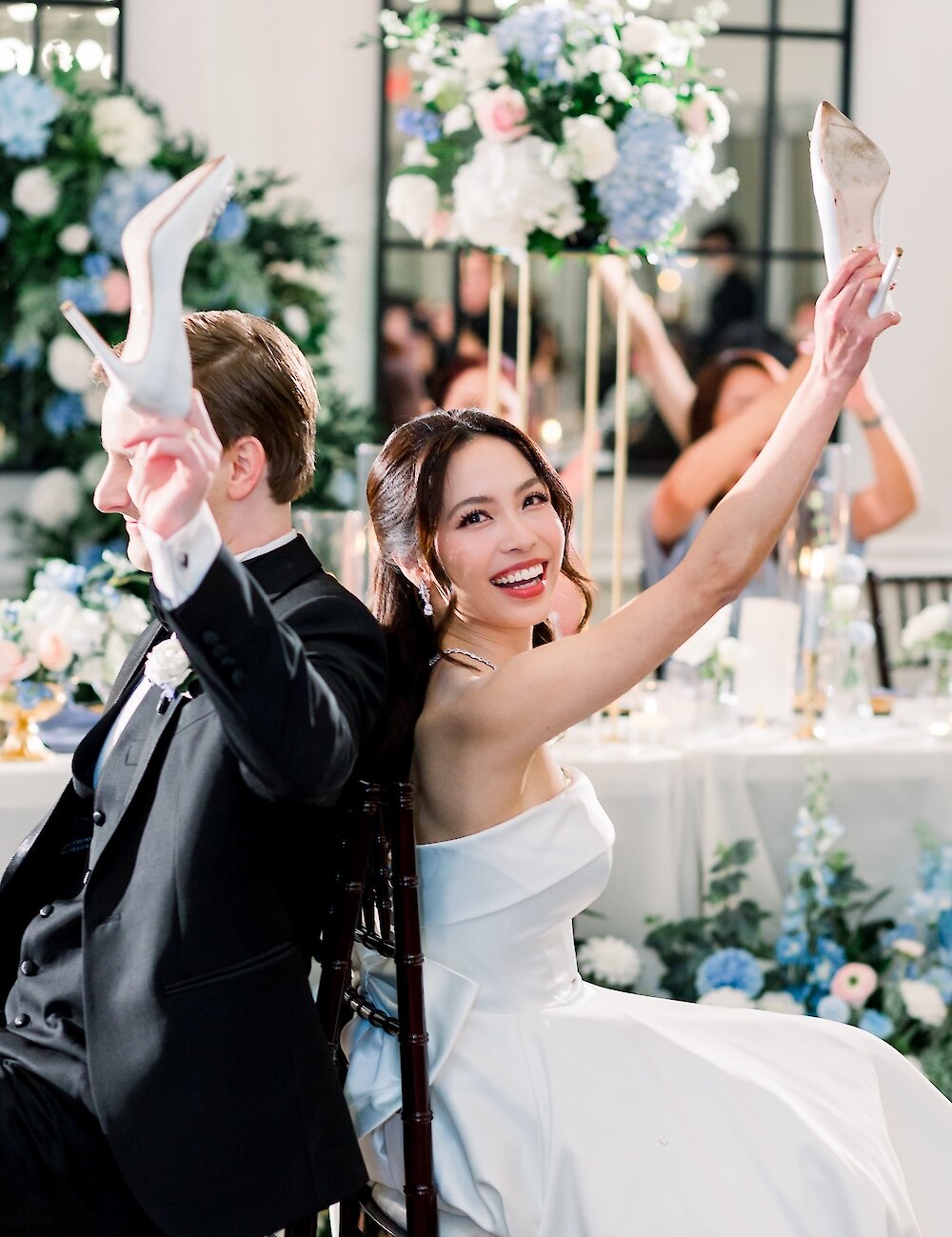 The bride and groom are sitting back-to-back, laughing and holding up their shoes during the popular wedding shoe game.