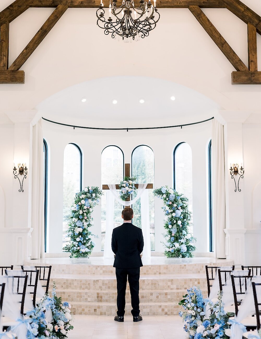 The groom stands facing a large wooden cross decorated with blue and white flowers in a bright, modern white chapel.