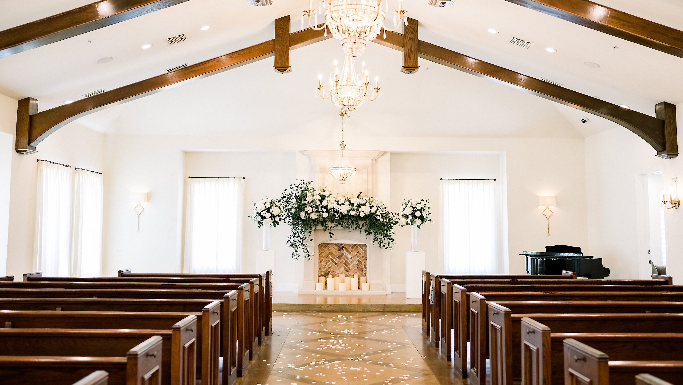 The interior of an elegant, traditional chapel featuring wooden pews, high ceilings, large chandeliers, and a white floral mantle.