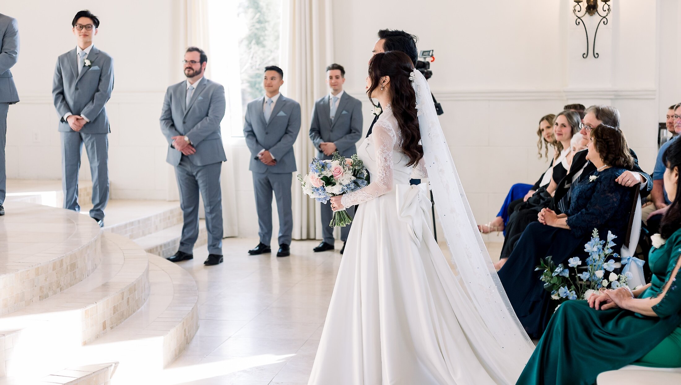 The bride looks toward the groom at the altar while holding a bouquet during the white and bright indoor wedding ceremony.