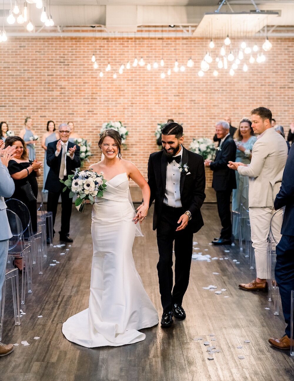 A joyful bride and groom exiting the ceremony aisle after their wedding, holding hands as guests applaud.